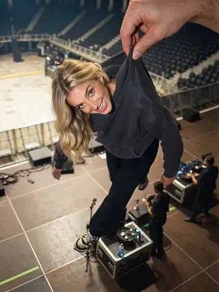 Tiny woman in a dark sweatshirt held up by giant fingers above an empty arena stage, laughing and kicking her legs